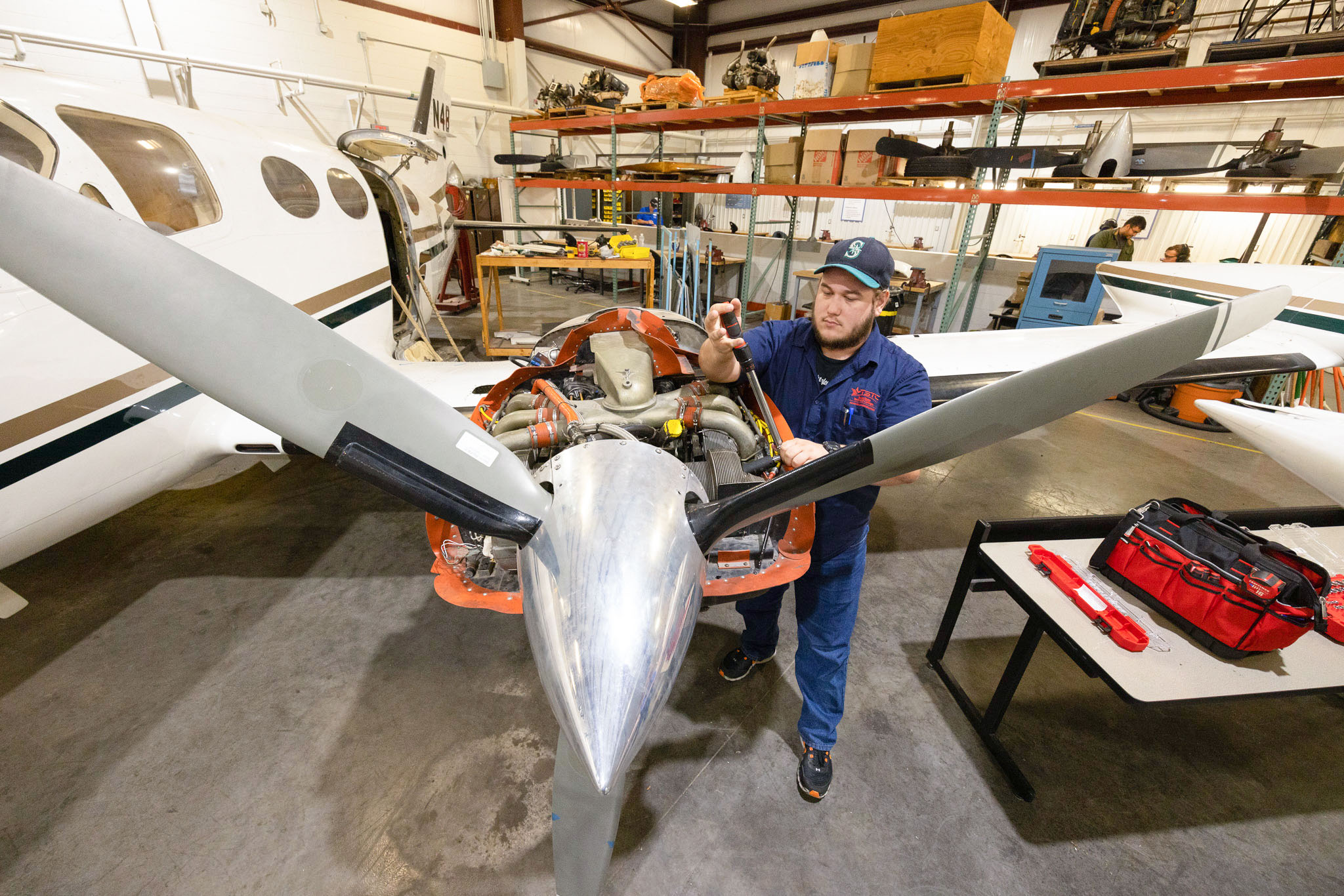 Student mechanic in a workshop uses a tool to work on an exposed aircraft engine mounted on a small propeller plane, with the propeller blades extending toward the camera and tools laid out nearby.